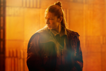 Hunter Schafer wearing a dark utilitarian jacket and bun, looking left in a glowing orange sci-fi interior.