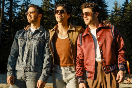 The Jonas Brothers in sunglasses and retro jackets stand outdoors, arms around each other, with a forest in the background.