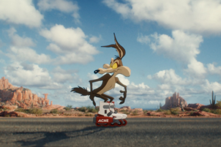 Wile E. Coyote skates on Acme roller skates on a desert road with mesas under a blue, cloudy sky.