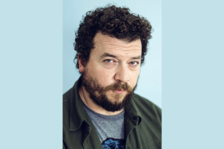 Man with curly dark hair and beard, wearing a dark green shirt, looks directly at the camera against a light blue background.