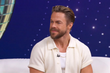 Derek Hough smiles, sitting on a white couch against a starry blue background with a disco ball.