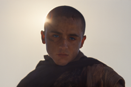 Young man with shaved head, blue eyes, and facial scars looks intently at viewer, backlit by a bright sun.