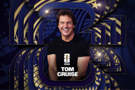 Tom Cruise smiles, wearing a black FIFA World Cup 2026 t-shirt with his name, against a blue and gold patterned background.
