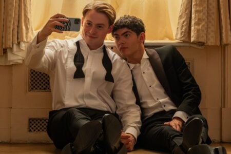 Two young men in tuxedos sit on the floor against a light wall, one smiling and taking a selfie, the other leaning on him.