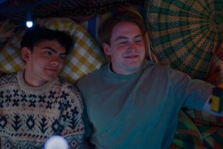 Two smiling men lie in a cozy fort of blankets and string lights, one holding a small yellow camera.
