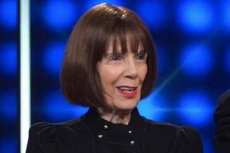 Woman with short brown hair, bangs, and red lipstick smiles, wearing a black jacket, against a blue studio background.