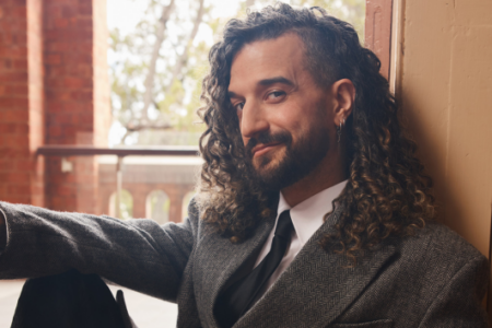 Man with long curly hair, beard, and suit jacket smiles, leaning against an indoor wall with a brick building outside.