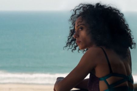 Woman with dark curly hair looks out from a balcony over a sandy beach and blue ocean.