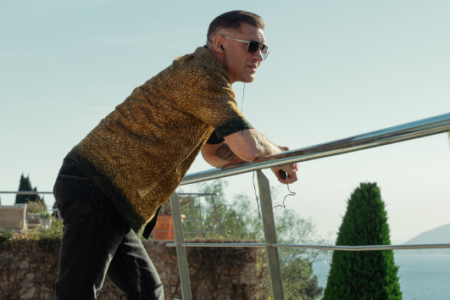 Man in patterned shirt, sunglasses, and earbuds leans on a railing overlooking a sunny body of water and hills.
