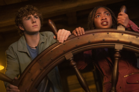 Inside a dim ship's cabin, a curly-haired white boy and Black girl grip a steering wheel, looking up with concern.