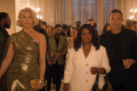 Blonde woman in gold, Octavia Spencer in white, and a man in black walk through a crowded, elegant party.