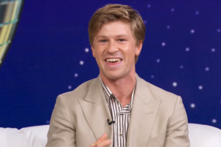 Robert Irwin laughs from a white couch against a blue, starry studio background with a disco ball.