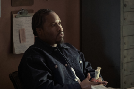 A Black man with braided hair sits in an office with a filing cabinet, holding stacks of cash with a red band.
