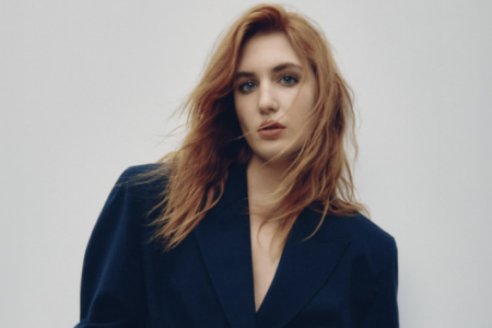 Reddish-blonde woman with blue eyes in a dark blue blazer against a plain light gray background.