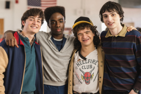 Four smiling Stranger Things actors pose arm-in-arm in a school hall, one in a "Hellfire Club" shirt.