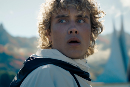 Surprised young man with curly blonde hair looks up, with mountains and modern spires in the background.