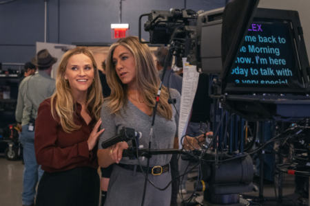 Reese Witherspoon smiles beside Jennifer Aniston, who focuses on a teleprompter attached to a studio camera.