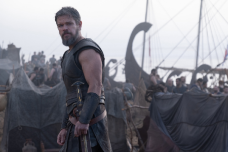 Bearded Matt Damon in warrior armor holds a sword, looking at viewer in a busy camp with ancient ships.