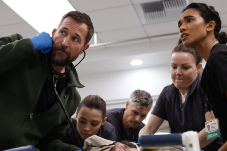 Medical team members, including a man with a stethoscope and a doctor, intently treat a patient on a gurney in an ER.