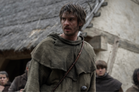 Young man with messy hair and beard in a torn hooded tunic looks right, in a medieval village with thatched buildings.