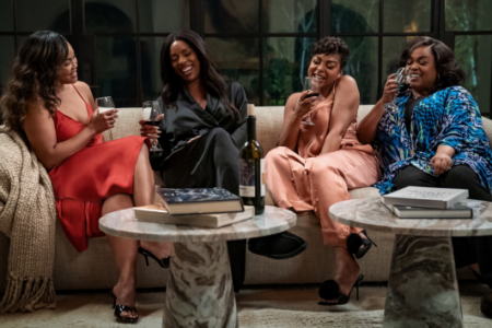 Four Black women laugh and drink wine on a sofa, with a wine bottle and books on coffee tables.