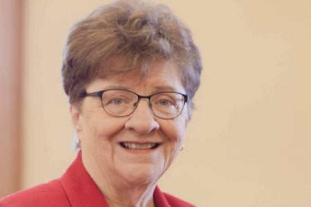 Older woman with short brown hair, dark-rimmed glasses, and a red blazer smiles warmly indoors.