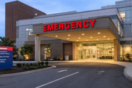 Brightly lit emergency entrance of a modern hospital at dusk, with a red 'EMERGENCY' sign and patient parking directions.