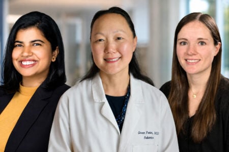 Three smiling women; the center woman wears a lab coat with "Grace Freier, M.D. Pediatrics," in a blurred clinic.