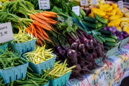 Fresh green beans, carrots, beets, cucumbers, and yellow squash are displayed at a vibrant farmers market.