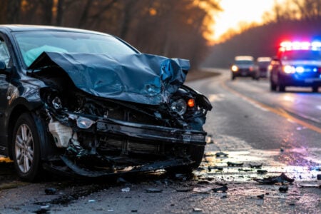 Front-end damaged dark car on a road with debris, and a police cruiser with flashing lights in the background.