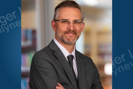 Man with glasses, beard, suit, and crossed arms smiles in front of blurred bookshelves.