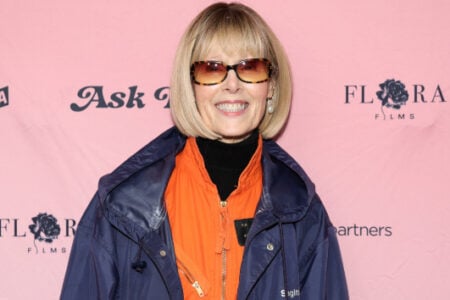 Woman with blonde bob and tortoiseshell sunglasses smiles in orange and blue hooded jacket against a pink media wall.