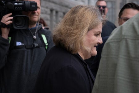 Blonde woman smiles while walking past a cameraman and other people outside a building.