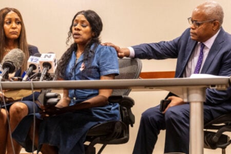 An emotional woman sits at a table with microphones, a man's hand on her shoulder, next to another woman, at a press conference.