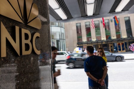 Gold NBC logo on a building, with people and cars on a busy street near Rockefeller Center in New York City.