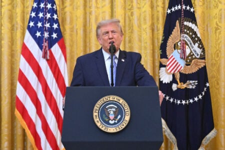 Donald Trump speaks at a White House press conference, flanked by U.S. and Presidential flags.