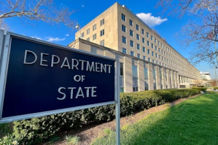 Department of State sign in front of its large building, surrounded by green landscaping under a clear blue sky.
