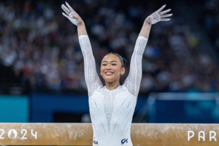 Smiling gymnast with chalky hands raised in celebration above a "PARIS 2024" balance beam.