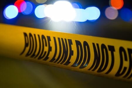 Close-up of yellow "POLICE LINE DO NOT CROSS" tape with blurred red, blue, and white police lights in the dark background.