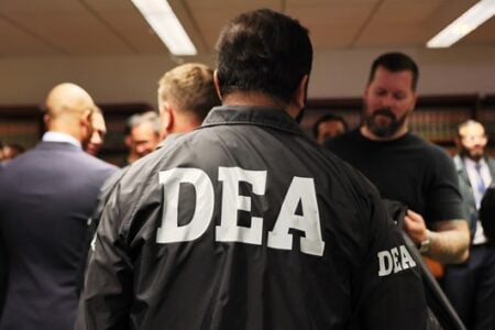 DEA agent in a black jacket with "DEA" on the back stands among people in a room with bookshelves.