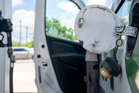Vehicle's open fuel door and dangling gas cap at a gas station pump.