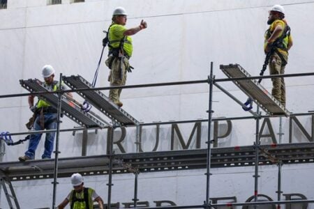 Construction workers on scaffolding next to large "TRUMP AN" letters on a building, one gives a thumbs up.