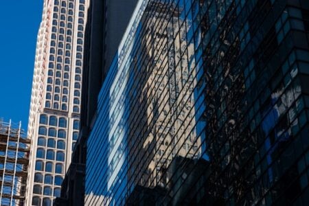 Glass skyscraper in an urban scene reflects a light-colored building and blue sky; scaffolding visible.
