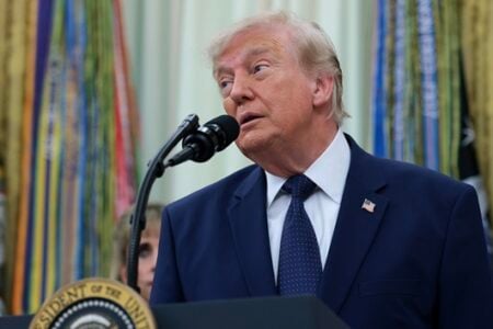Donald Trump speaking at a podium with the Presidential Seal, wearing a blue suit and tie, colorful drapes in background.
