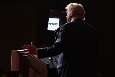 Donald Trump speaks from a podium on a dark stage, gesturing, with "sovereignty" visible on his teleprompter.