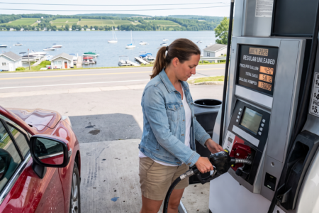 Woman pumping gas into a red car at a lakeside gas station, with boats on the lake and hills in the background.