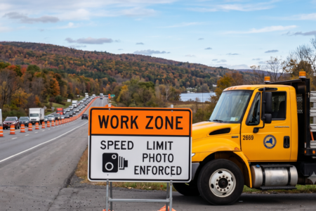 Yellow NY DOT truck and "Work Zone Speed Limit Photo Enforced" sign on an autumn highway with traffic.