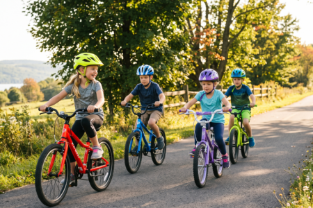 Four happy children wearing helmets ride bikes down a paved path lined with trees and wildflowers, with a lake in the distance.
