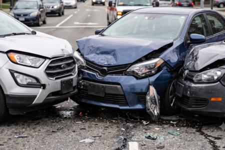 Three vehicles, a silver SUV, blue sedan, and gray car, severely damaged in a collision on a city street, with debris.