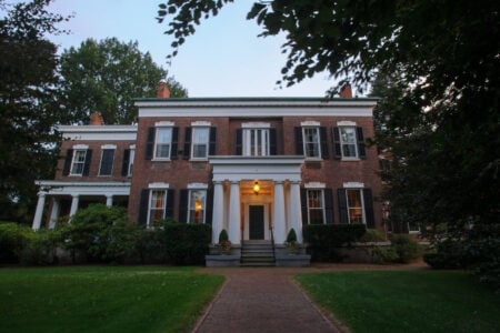Large brick house with white columned portico, illuminated entrance, brick path, and lawn at dusk.